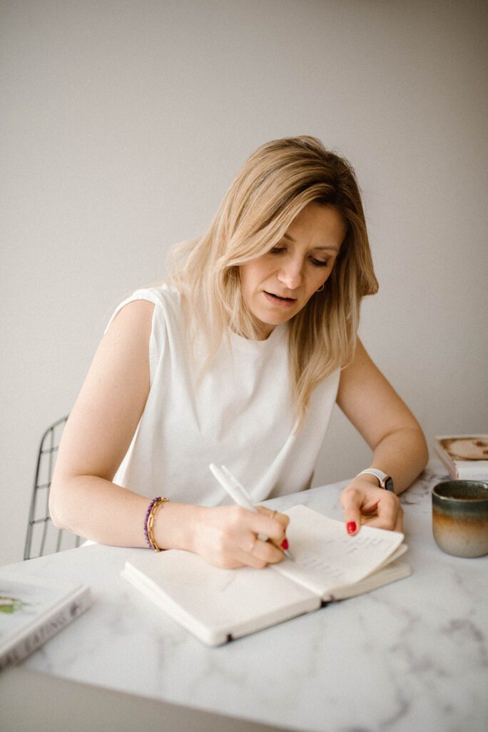 a woman in white t-shirt sitting at a desk and writing in a notebook, with her head and hair down